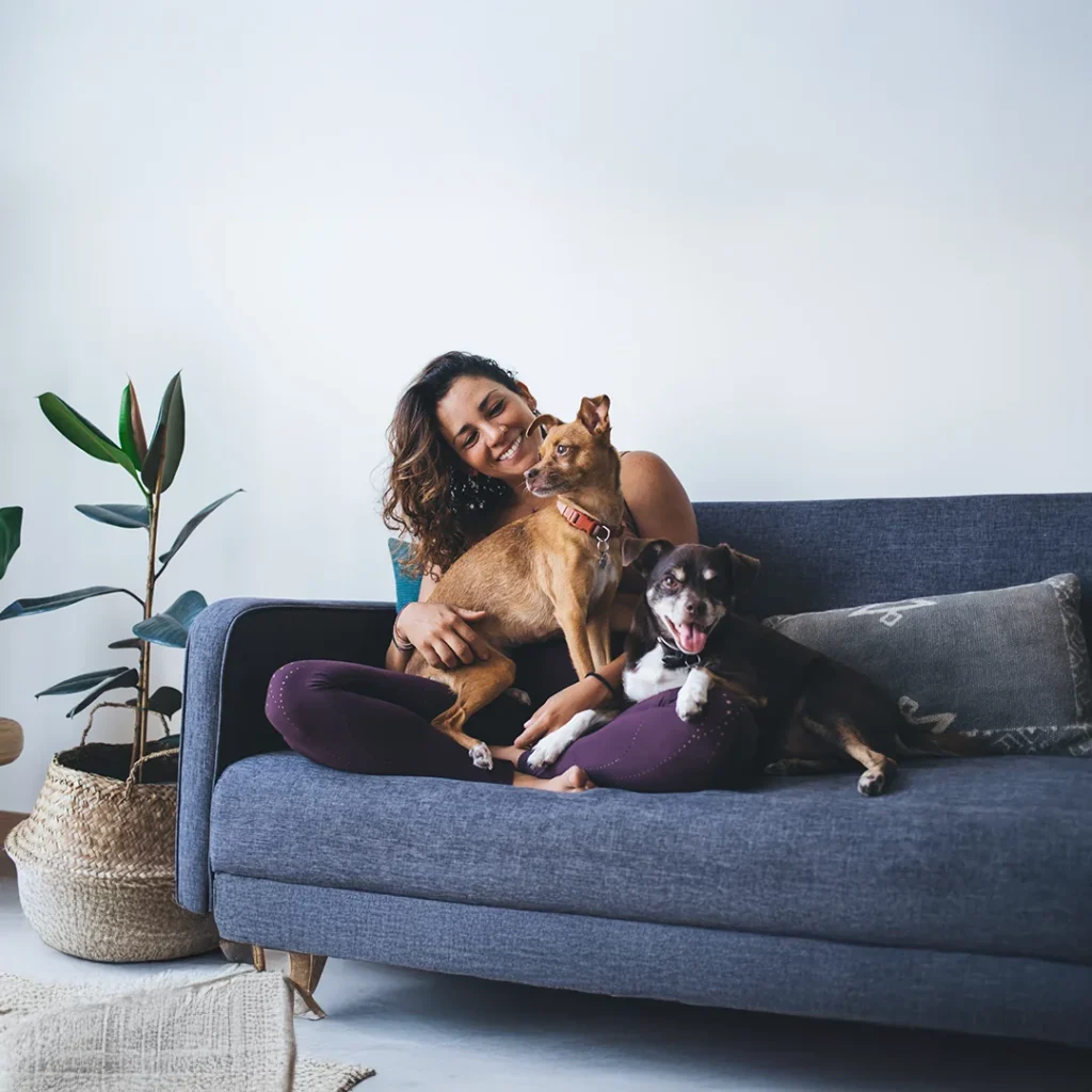 Pet Owner Relaxing With Their Dogs at Home Person sitting on a couch smiling while cuddling two dogs in a calm, comfortable living room